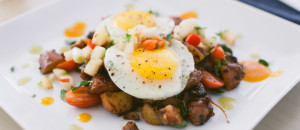 white plate with potatos, grape tomatoes, green herbs and orange hot sauce topped with two fried eggs