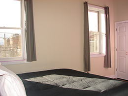 king bed with white pillows and sunlight hitting the bed from two windows with grey curtains.