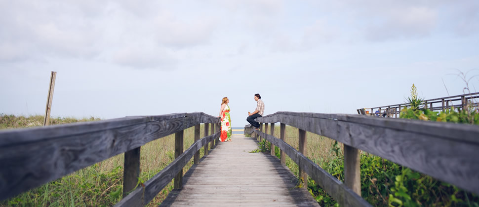 Heather in long flowing dress with red and green print across from Mike sitting on the wooden walkway to the beach