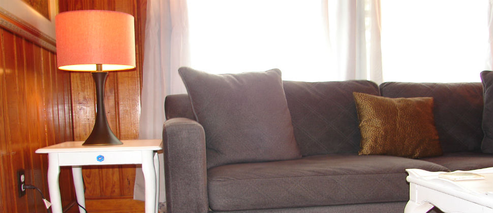 living room, blue/grey couch in front of two windows, white end table with illuminated brown lamp and orange shade.