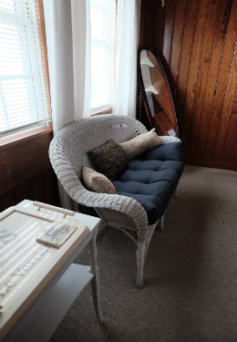 White wicker loveseat with a blue cusion against two windows with white drapes and antique mirror against the back wall