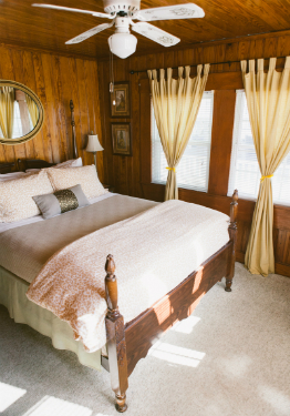 Queen bed with tan blanket and tan and white duvet. Two windows with yellow drapes and white blinds.