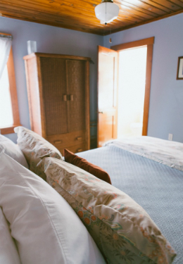 Queen bed with blue blanket and tan pillow shams. Brown wicker hutch in background with open door leading to bathroom.