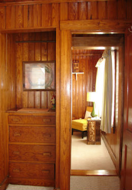 Honey stained knotty pine walls with built in dresser, peaking through to bedroom with yellow chair and green lamp.