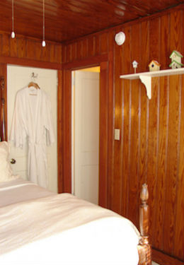 Bed with duvet folded at the end of the bed, white bathrobes hanging on the door, honey stained knotty pine walls.