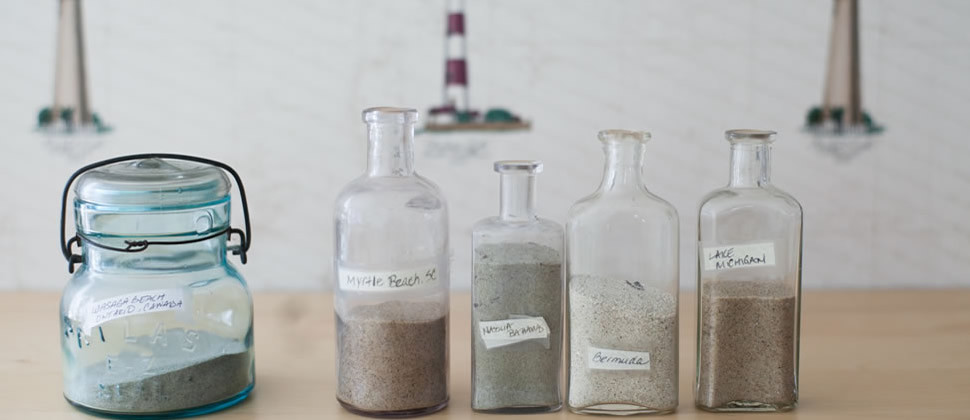 five old medicine bottles of sand from various beaches set on tan wooden table with lighthouse wallpaper in background