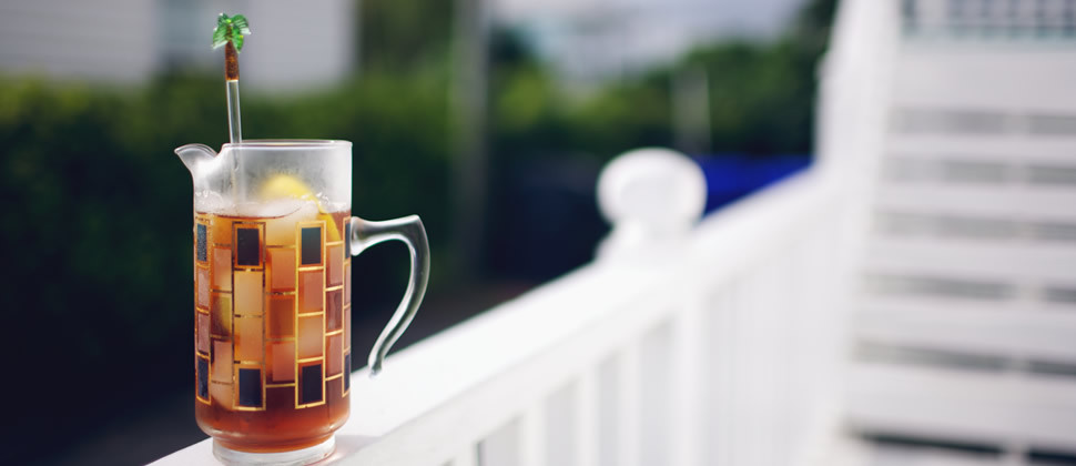 ice tea in clear glass decorated with black and gold piece of yellow lemon and palm tree stirrer sitting on white ledge of back deck